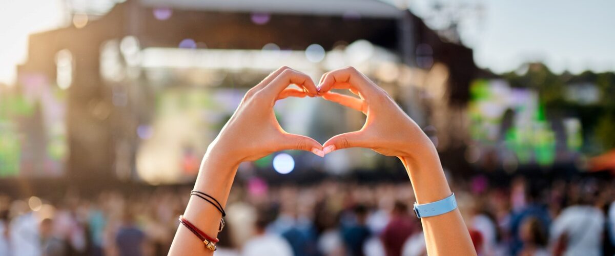 A concertgoer makes a heart shape with their hands at an outdoor music festival during the day.