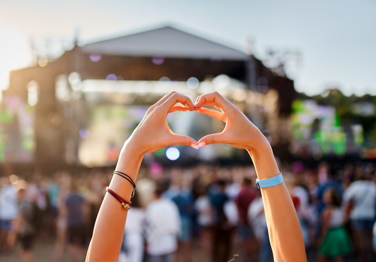 A concertgoer makes a heart shape with their hands at an outdoor music festival during the day.