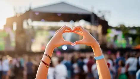 A concertgoer makes a heart shape with their hands at an outdoor music festival during the day.