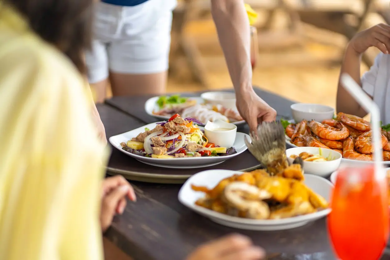 A server places plates of seafood onto a wooden table for customers at a beachside restaurant. 
