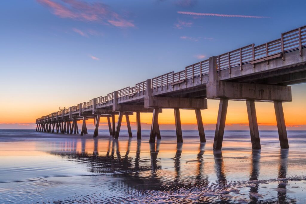 The Jacksonville Beach Pier stretching over calm ocean waters at sunrise, reflecting colors on the wet sand below.