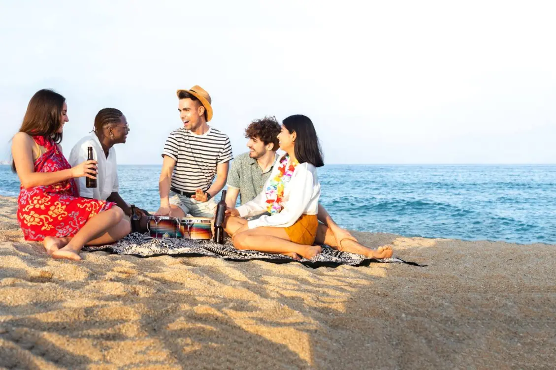 A group of friends sitting on a beach blanket, playing music and enjoying drinks together by the ocean on a sunny day.