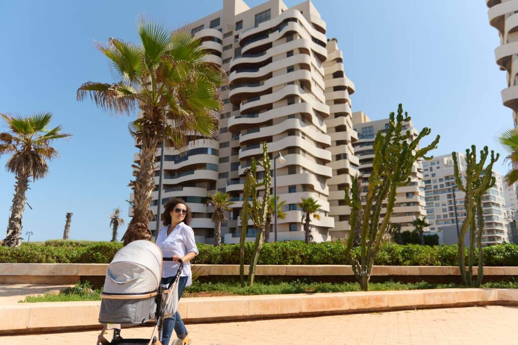 A woman pushes a stroller along a sunny walkway lined with palm trees and cacti, and modern high-rise buildings behind.
