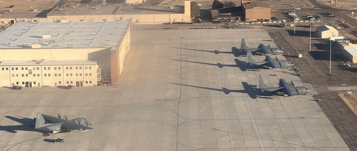 Several military transport aircraft parked on a large concrete airfield near hangars at a naval air force base.