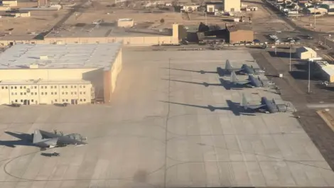 Several military transport aircraft parked on a large concrete airfield near hangars at a naval air force base.
