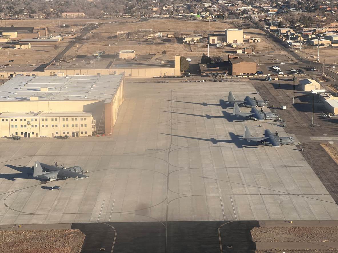 Several military transport aircraft parked on a large concrete airfield near hangars at a naval air force base.