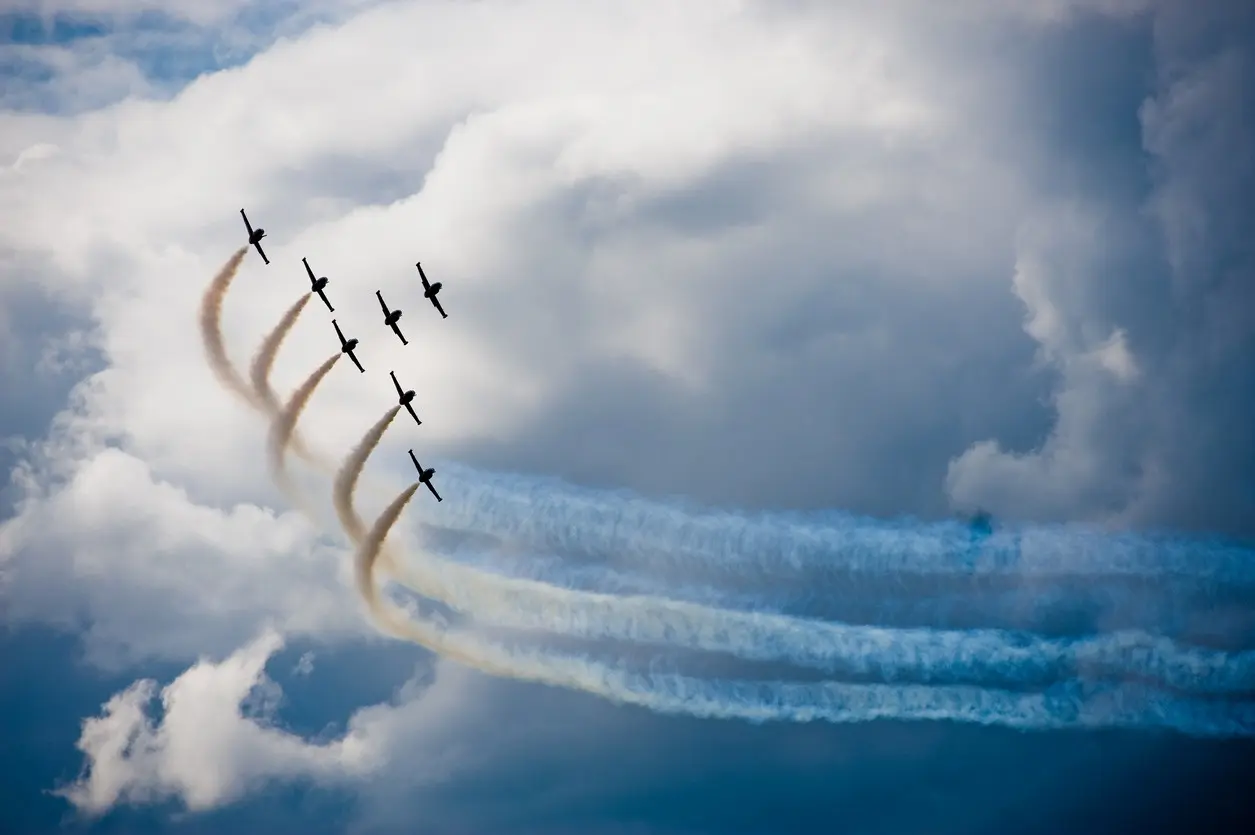 A group of planes performs a synchronized air show with colorful smoke trails against a cloudy sky.