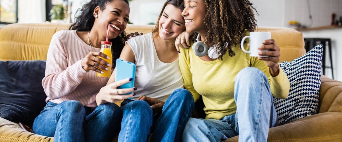 Three young women sitting on a couch, laughing and taking a selfie together, and enjoying drinks in their apartment.