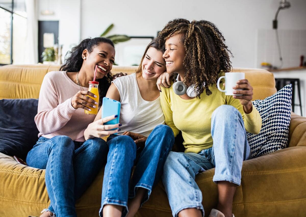 Three young women sitting on a couch, laughing and taking a selfie together, and enjoying drinks in their apartment.