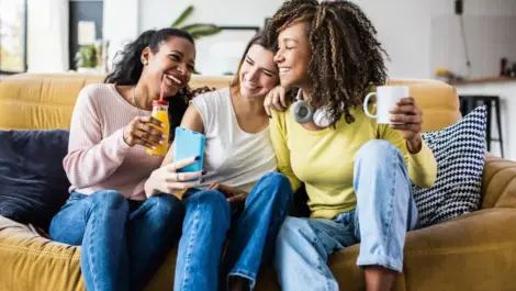 Three young women sitting on a couch, laughing and taking a selfie together, and enjoying drinks in their apartment.