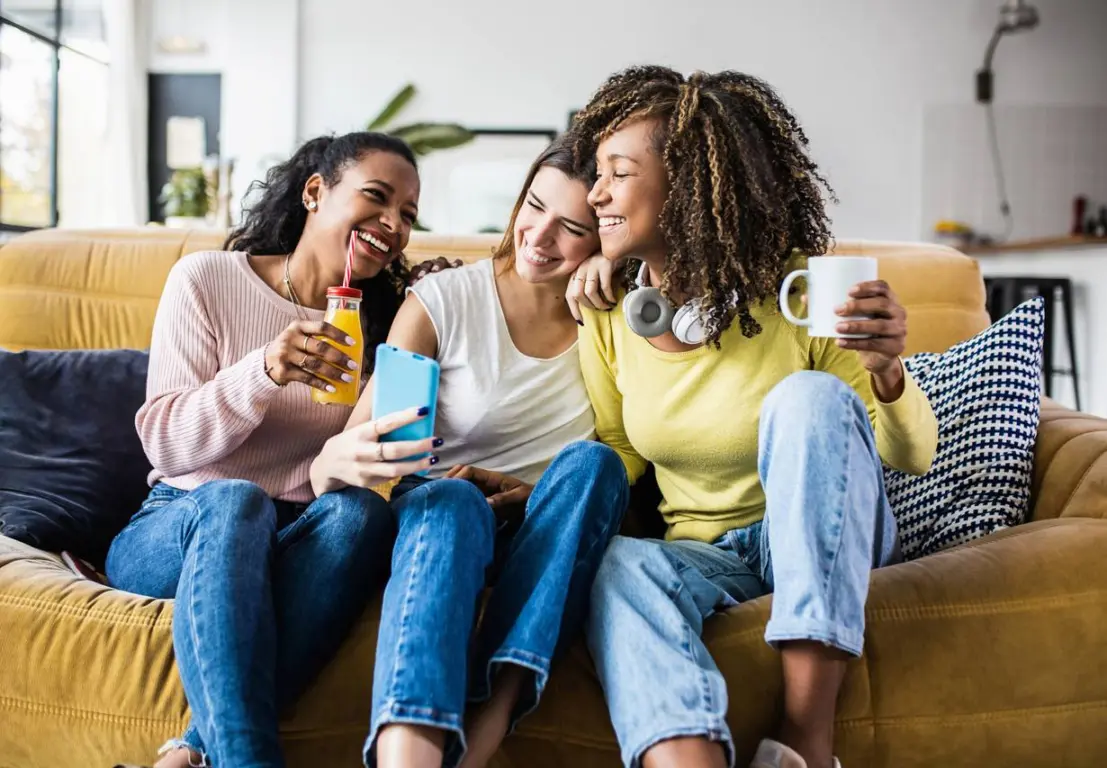 Three young women sitting on a couch, laughing and taking a selfie together, and enjoying drinks in their apartment.