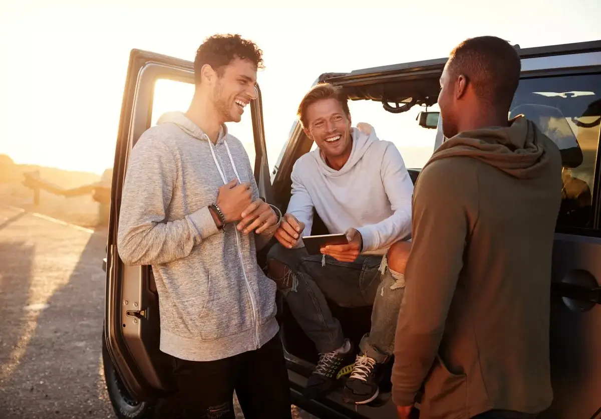 Three friends laugh and talk beside a parked car at sunset, enjoying a break together during their road trip.