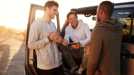 Three friends laugh and talk beside a parked car at sunset, enjoying a break together during their road trip.