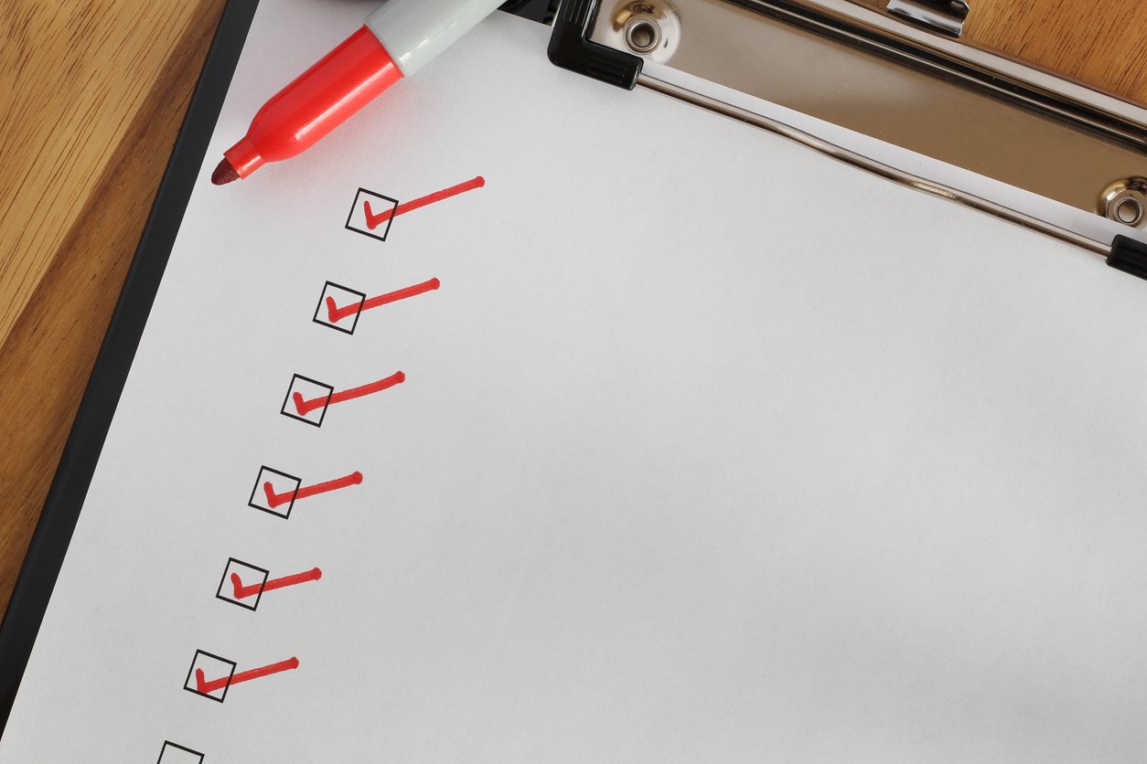 A red marker rests on a clipboard beside a checklist with several boxes marked with red checkmarks on a wooden desk.