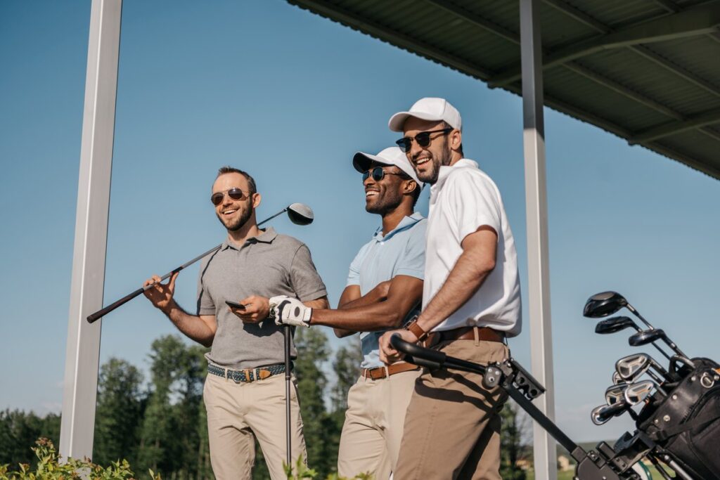 Three golfers laugh and talk under a covered golf area, holding clubs and standing beside a golf bag on a sunny day. 