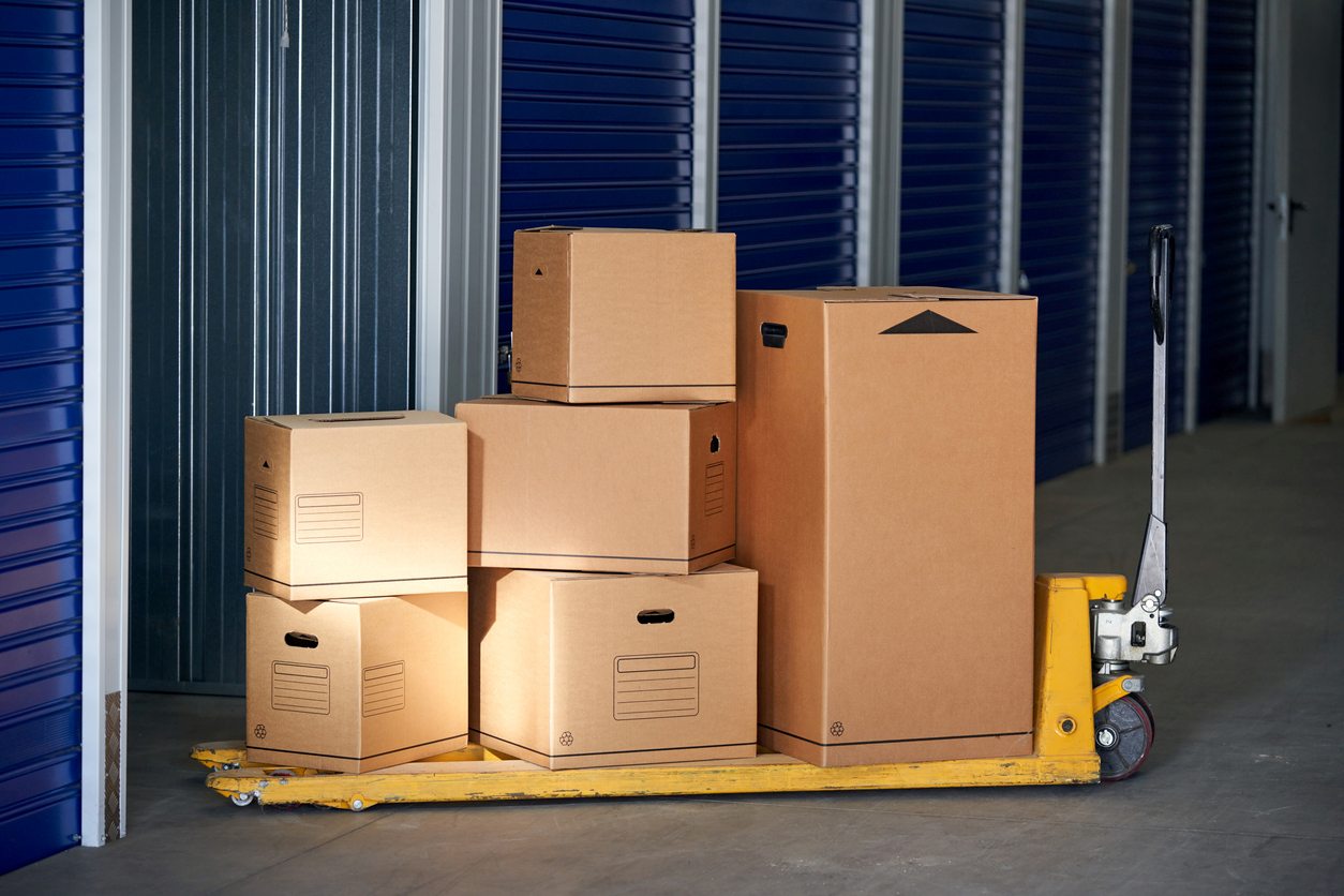 A stack of moving boxes on a yellow pallet jack inside a storage facility hallway with blue doors. 