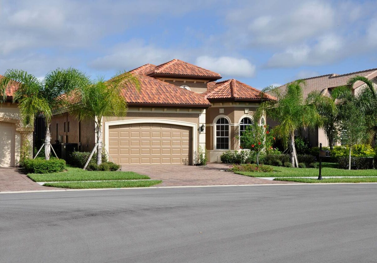 A luxury Florida-style home with a tile roof, palm trees, and a clean driveway on a sunny day in a neighborhood.