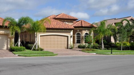 A luxury Florida-style home with a tile roof, palm trees, and a clean driveway on a sunny day in a neighborhood.