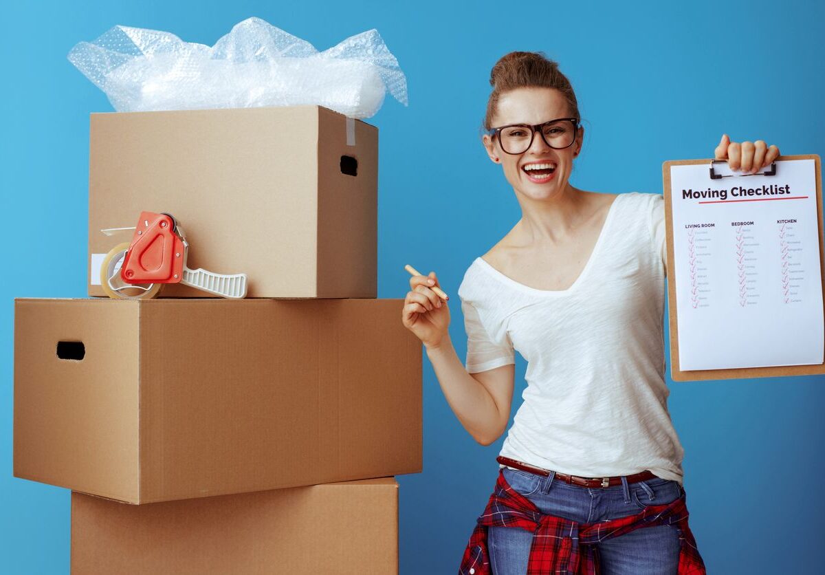 A smiling woman holding a moving checklist beside cardboard boxes.