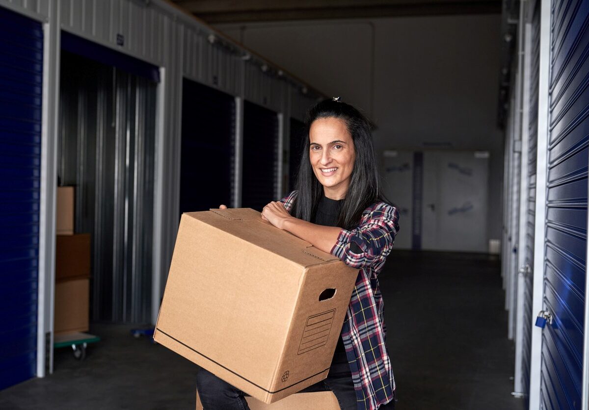 Woman with a heavy cardboard box smiling while sitting near self storage during moving day.