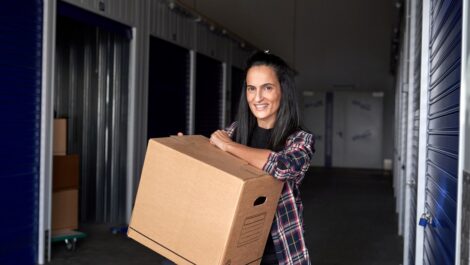 Woman with a heavy cardboard box smiling while sitting near self storage during moving day.